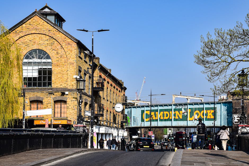 A busy street scene in Camden, London, featuring a two-storey brick building with large arched windows on the upper level and smaller rectangular windows below, adjacent to a railway bridge with a sign displaying 'Camden Lock' in yellow letters against a blue background. Several pedestrians walk along the sidewalk, some carrying bags, with vehicles including cars and a bus visible on the road. A street clock is mounted on a pole near the building, and a trolleybus overhead wire system stretches across the scene. The environment is lively, with trees partially visible on the right side and a clear blue sky overhead. This setting relates to house removals and urban moving logistics, highlighting furniture transport, packing, and loading processes typical of home relocations facilitated by Kentishtown Removals.