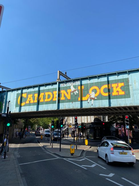 A blue metal railway bridge with the words 'Camden Lock' painted in large yellow letters spans above a busy street in Camden, London. The bridge features some graffiti and a small security camera. Below the bridge, cars, including a white vehicle, are driving through traffic lights, while pedestrians walk on the sidewalks and cross the road. Orange and black bollards, traffic signs, and a pedestrian crossing are visible along the street. The setting suggests an urban environment involved in home relocation or furniture transport, with Kentishtown Removals possibly managing moving logistics in this area.
