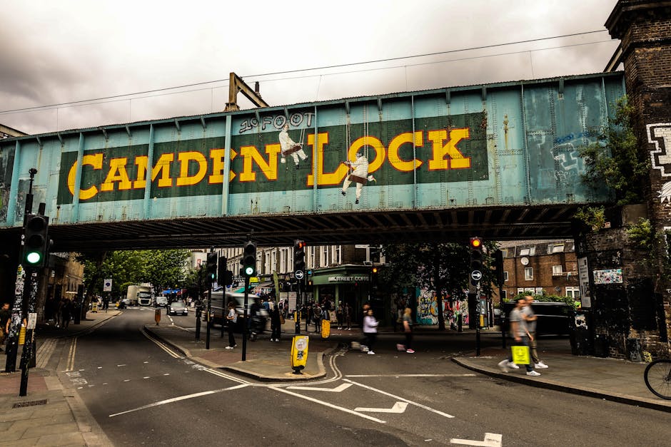 A large, weathered green metal railway bridge with the words 'CAMDEN LOCK' painted in bold yellow letters across the side. The bridge is situated above a busy street with pedestrians crossing at the curb and traffic signals controlling vehicle flow. Several individuals are walking on the pavement, some carrying backpacks or bags, while others are waiting to cross or walking in different directions. Visible behind the bridge are buildings with brick exteriors, some covered in graffiti, indicating an urban environment. The scene includes various items associated with house removals, such as large cardboard boxes, moving blankets, and reusable plastic packaging, suggesting a home relocation process. The natural lighting is overcast, typical of a cloudy day, and the setting supports the logistics involved in furniture transport and packing during house removals in Camden.