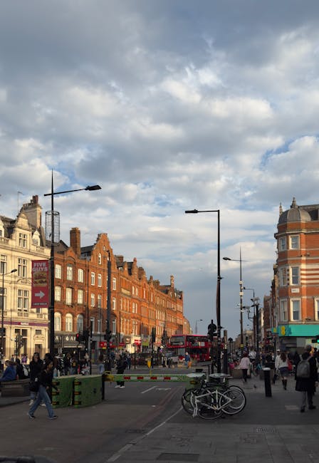 A busy street scene in Camden, London, with a backdrop of historic red brick buildings and a prominent corner structure featuring a rounded tower with a decorative roof. The street is active with pedestrians walking on the pavement and crossing the road, some carrying shopping bags. In the foreground, there are green and black wheelie bins, bicycles parked near a barrier, and street furniture including lampposts and traffic lights. Overhead, a partly cloudy sky is visible, with some blue sky peeking through. Visibility of loading or moving activities is limited, but the setting reflects an urban environment suitable for house removals and furniture transport, as addressed by Kentishtown Removals, especially in relation to Camden Council permit rules for relocations.