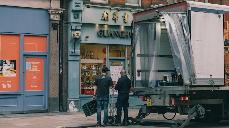 Two movers from Kentishtown Removals are preparing to load a delivery truck outside a storefront on a city street, which has signage with Chinese characters and the word 'GUANGH.' One mover, dressed in dark clothing, holds a crate, while the other, also in dark attire, is standing nearby with a dolly. The truck's rear door is open and covered with a transparent plastic sheet, revealing empty interior space suitable for transporting furniture or packed boxes during home relocation or furniture transport. The sidewalk features a grey paving surface, and adjacent buildings have decorative architectural elements, with a blue facade and orange panels. This scene captures the loading process involved in packing and moving services, illustrating the careful handling of items in an urban environment, aligned with the context of delays that affect Kentish Town removals, as discussed on kentishtownremovals.co.uk.