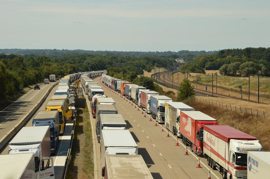 A long line of trucks and lorries parked on the side of a dual carriageway during daytime, with some vehicles partially obstructing the traffic lane. The trucks vary in size, color, and branding, with some carrying visible cargo containers and others with empty cargo areas. Traffic cones are placed along the edge of the lane to manage the queue, and there is a clear division between the parked trucks and the moving traffic lane on the left side of the image. The background features open fields, grassy areas, and distant trees, with overhead power lines running parallel to the road. The weather appears to be partly cloudy, with sufficient natural light illuminating the scene. This image captures a waiting or delay scenario that might occur during home relocation or furniture transport logistics, highlighting the importance of planning for potential transportation delays during house removals, as discussed on the page about delays affecting Kentish Town removals by Kentishtown Removals.