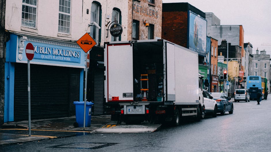 Two movers from Kentishtown Removals are preparing to load a delivery truck outside a storefront on a city street, which has signage with Chinese characters and the word 'GUANGH.' One mover, dressed in dark clothing, holds a crate, while the other, also in dark attire, is standing nearby with a dolly. The truck's rear door is open and covered with a transparent plastic sheet, revealing empty interior space suitable for transporting furniture or packed boxes during home relocation or furniture transport. The sidewalk features a grey paving surface, and adjacent buildings have decorative architectural elements, with a blue facade and orange panels. This scene captures the loading process involved in packing and moving services, illustrating the careful handling of items in an urban environment, aligned with the context of delays that affect Kentish Town removals, as discussed on kentishtownremovals.co.uk.