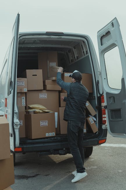 A person wearing a dark jacket, matching trousers, a baseball cap, and white sneakers is inside the open back of a large grey van, stacking and managing labelled cardboard boxes during a home relocation. The boxes are of various sizes, some sealed with packing tape, and are arranged on the van's loading area and the driveway adjacent to the property. The individual is holding a box, preparing to load or unload it, with the van situated outdoors under natural lighting. Behind them, the vehicle doors are wide open, revealing the interior space filled with several packed boxes, some foam padding, and protective materials used for furniture transport and packing during a moving process. This scene depicts the loading process typical of furniture transport and home removals, with a focus on efficient logistics for difficult access moves, as handled by Kentishtown Removals.