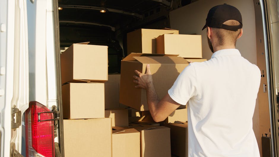A man wearing a white t-shirt, cap, and glasses is inside a moving van, packing and loading medium-sized cardboard boxes onto the vehicle. The boxes are stacked neatly, some with open flaps, and are made of brown corrugated cardboard with visible packing tape. The van's interior is dimly lit, with the open rear doors revealing a spacious loading area filled with packing materials such as plastic wrap and bubble wrap, indicating careful preparation for a home relocation. Outside the van, part of the pavement and the vehicle's exterior can be seen, suggesting the loading process is taking place outdoors, possibly on a residential street or driveway. A professional removal service like Kentishtown Removals specializes in difficult access moves in Kentish Town, ensuring furniture and belongings are transported safely and efficiently.