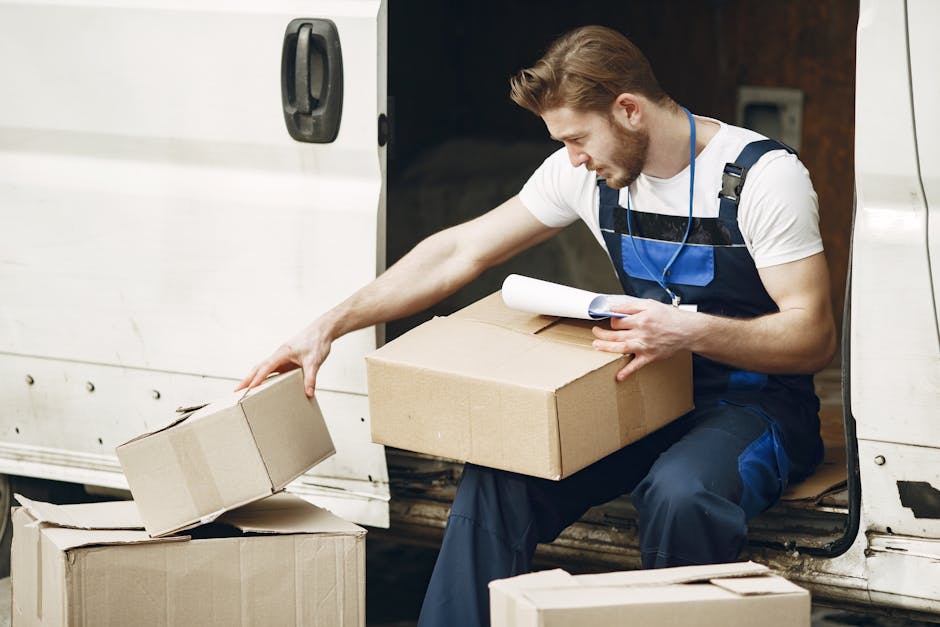 A person wearing a dark jacket, matching trousers, a baseball cap, and white sneakers is inside the open back of a large grey van, stacking and managing labelled cardboard boxes during a home relocation. The boxes are of various sizes, some sealed with packing tape, and are arranged on the van's loading area and the driveway adjacent to the property. The individual is holding a box, preparing to load or unload it, with the van situated outdoors under natural lighting. Behind them, the vehicle doors are wide open, revealing the interior space filled with several packed boxes, some foam padding, and protective materials used for furniture transport and packing during a moving process. This scene depicts the loading process typical of furniture transport and home removals, with a focus on efficient logistics for difficult access moves, as handled by Kentishtown Removals.