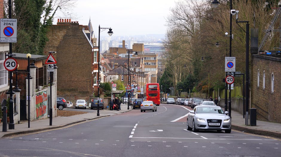 A street scene showing a residential area with brick and stucco buildings on both sides, featuring various road signs including a controlled zone and 20 mph speed limit. A sidewalk runs along the left side, where a few people are waiting near a bus stop. In the foreground, a silver Audi car is parked partially on the pavement, and further back, a grey vehicle is driving along the curved road. A red double-decker bus is approaching from the distance, indicating bus services typical in urban home relocation routes. Street lamps, trees, and parked cars line the street, while a construction crane is visible on the right side, suggesting possible building activity nearby. The scene captures typical urban planning elements relevant to managing vehicle and pedestrian movement during house removals and furniture transport in Kentish Town, with the image illustrating the environment where Kentishtown Removals might operate during relocation logistics, including loading or unloading activities in constrained street access conditions.
