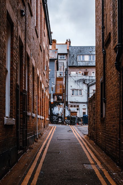 A narrow alleyway between two brick buildings in an urban area, with a paved surface featuring double yellow lines along the edges. At the far end of the alley, a small stack of cardboard boxes and plastic-wrapped furniture are visible, indicating recent packing or loading activity. Behind the boxes, there are black waste bins and a large, weathered grey building with multiple windows and external air conditioning units. One building has a fire escape staircase attached to its exterior, leading upstairs. The sky above is overcast, casting diffuse lighting over the scene, which appears to be part of a home relocation or furniture transport process managed by Kentishtown Removals, as suggested by the context of the page about Fortess Road access tips for Kentish Town removals.