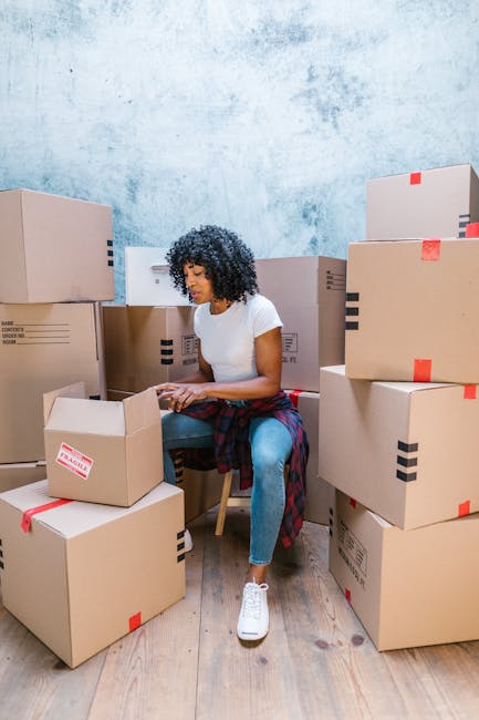 A woman with curly black hair, wearing a white t-shirt, blue jeans, and white sneakers, is seated among numerous cardboard boxes of various sizes, some with black and red packing tape, inside a room with a textured light blue wall and wooden flooring. She is holding and opening a cardboard box, surrounded by the boxes arranged in clusters on the floor, with some stacked on top of each other. The boxes appear to contain household items or belongings prepared for a home relocation. In the background, a partially visible door frame suggests this is an interior space being organized for moving. The scene depicts the packing process as part of furniture transport or house removals, with a focus on loading and packing tasks managed by Kentishtown Removals, a professional removals company.