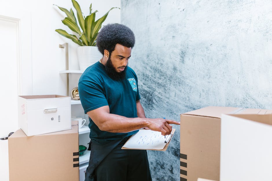 A woman with curly black hair, wearing a white t-shirt, blue jeans, and white sneakers, is seated among numerous cardboard boxes of various sizes, some with black and red packing tape, inside a room with a textured light blue wall and wooden flooring. She is holding and opening a cardboard box, surrounded by the boxes arranged in clusters on the floor, with some stacked on top of each other. The boxes appear to contain household items or belongings prepared for a home relocation. In the background, a partially visible door frame suggests this is an interior space being organized for moving. The scene depicts the packing process as part of furniture transport or house removals, with a focus on loading and packing tasks managed by Kentishtown Removals, a professional removals company.