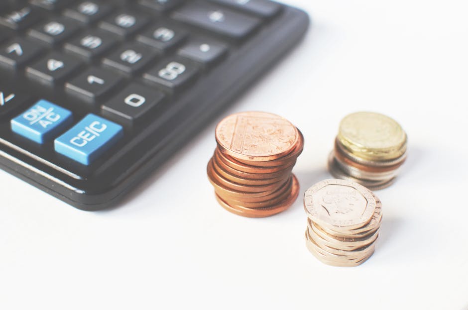 A close-up image showing a black calculator with blue and white buttons placed on a white surface, alongside three separate stacks of UK coins consisting of copper, silver, and gold coloured coins. Th