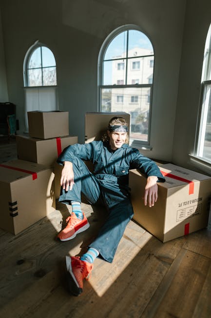 An individual dressed in blue work attire, including a cap and sneakers, sitting on a wooden floor surrounded by several cardboard boxes sealed with red and black tape inside a room with large arched windows. The windows let in natural daylight, illuminating the space and showing a view of neighboring buildings and trees outside. The boxes are of various sizes, some placed directly on the floor, while others are stacked. The scene depicts a home relocation or furniture transport process, likely involving packing and moving activities typical of professional removals services. The environment appears clean, well-lit, and prepared for the next stage of the move, representing the logistics and packing part of a house removal carried out by Kentishtown Removals.