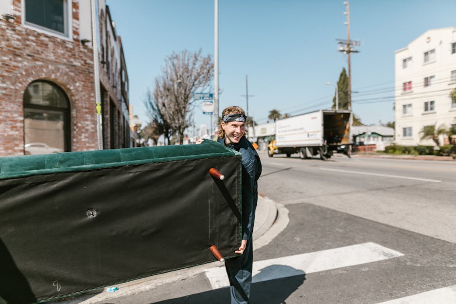 A man wearing a black jacket and a patterned headband is walking on a city street while carrying a large, padded furniture moving case with wooden handles, partially wrapped in green fabric padding. The background shows an intersection with a moving truck parked on the curb, its cargo area open, as well as multi-storey residential buildings, leafless trees, and utility poles with power lines. The scene appears to be part of a home relocation process, with the man possibly involved in furniture transport or packing and moving activities. The environment is bright and clear, with a blue sky and urban surroundings, indicating a typical day for local removals services such as Kentishtown Removals, which specialises in house removals and relocation logistics in NW5. The image conveys the practical aspects of furniture handling and loading procedures during a professional move.