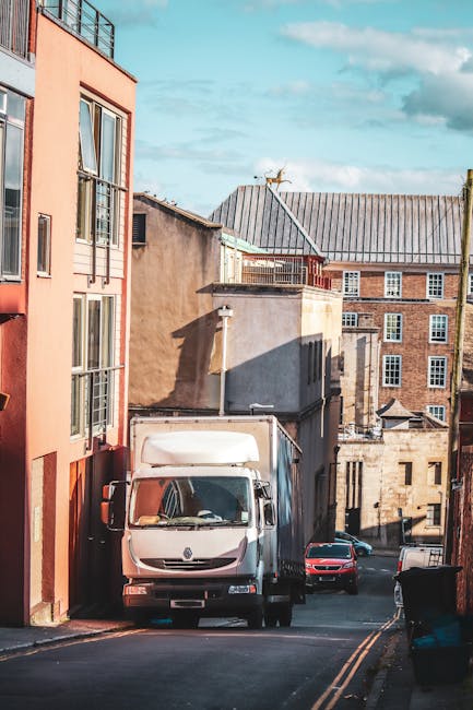 A white moving van with a closed cargo area is parked on a narrow, sloped street in an urban residential area during daylight hours. The van is positioned next to a pinkish-orange apartment building with large windows and balconies visible on the left side of the image. Behind the van, there are various older brick and concrete buildings with flat and sloped roofs, some featuring chimneys and aerials. The street includes a black wheelie bin on the right pavement and features yellow dashed lines indicating parking restrictions. Bright sunlight casts shadows of the buildings and the van onto the street, suggesting clear weather. The scene captures the typical environment for home relocation or furniture transport, with elements consistent with house removals services in Kentish Town, as provided by Kentishtown Removals, which specializes in move logistics and packing support.