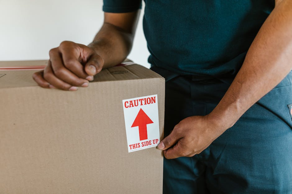 Two professional movers from Kentishtown Removals, dressed in navy uniforms, are engaged in a furniture loading process at the back of a white van. One mover is holding the side of a large, dark green upholstered piece of furniture, likely a sofa or armchair, while the other stabilizes it from the opposite side. The van's open rear doors reveal an interior with additional packing materials, possibly blankets or fabric wraps, used to protect the furniture during transportation. The scene takes place outdoors on a paved street under daylight, with a street sign and some other vehicles visible in the background. The equipment includes a trolley or dolly visible near the van, emphasizing careful handling. The image illustrates the logistics involved in home relocation, specifically furniture transport and packing within a moving or removals service, aligned with the services provided by Kentishtown Removals as highlighted on their website about same day removals in Kentish Town.